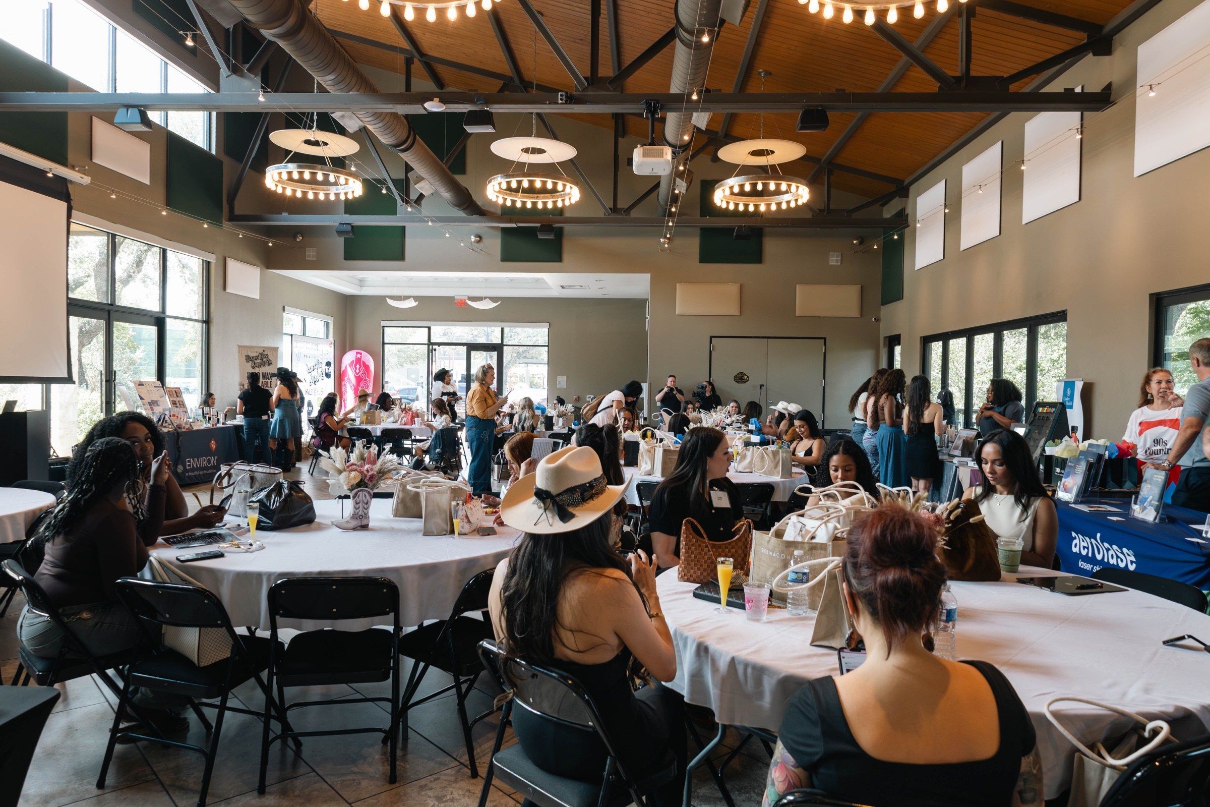A wide view of the Glow & Gather event room filled with attendees at round tables beneath circular chandeliers