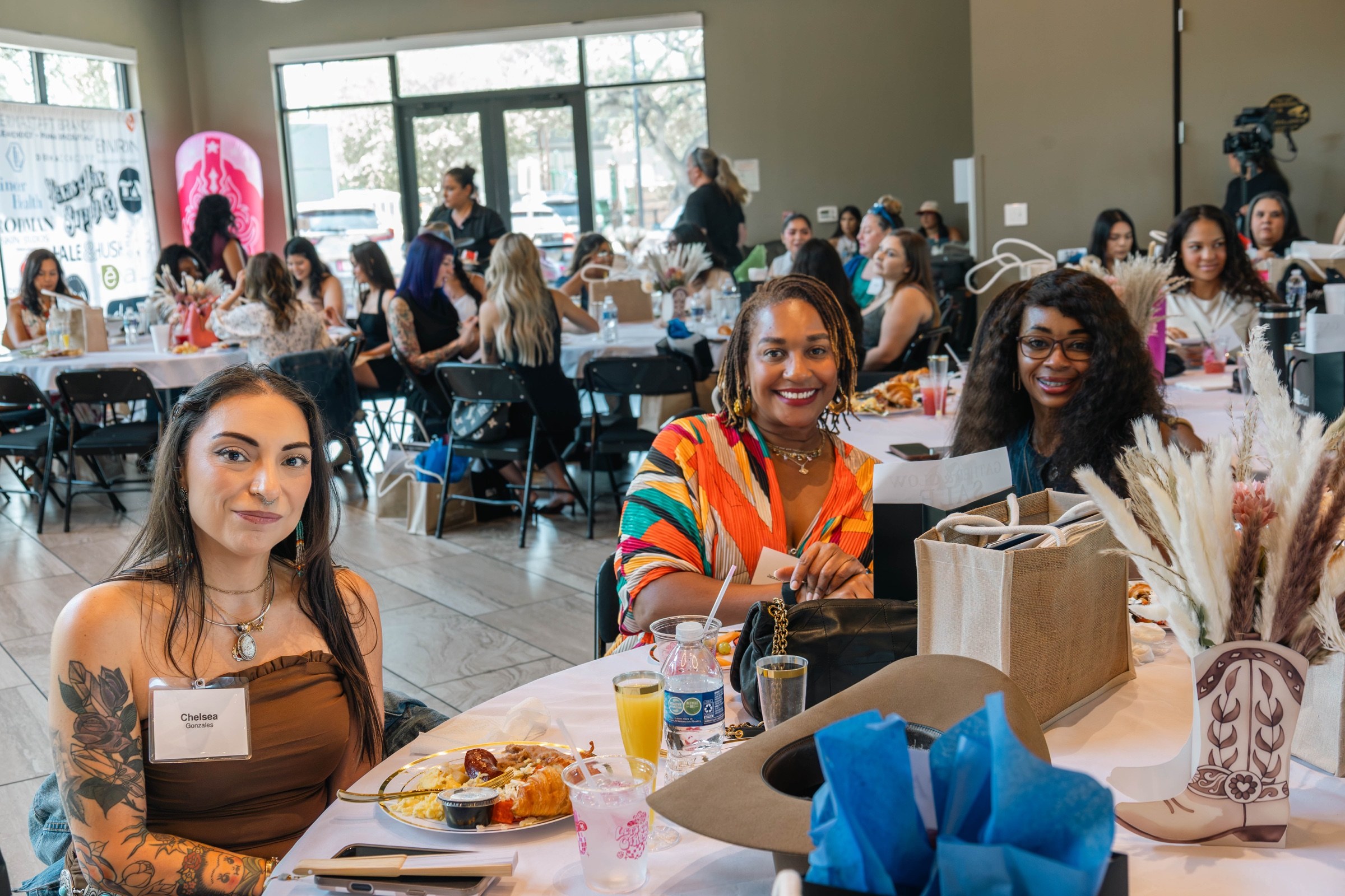 Three smiling attendees gathered at a decorated table with the busy Glow & Gather room behind them